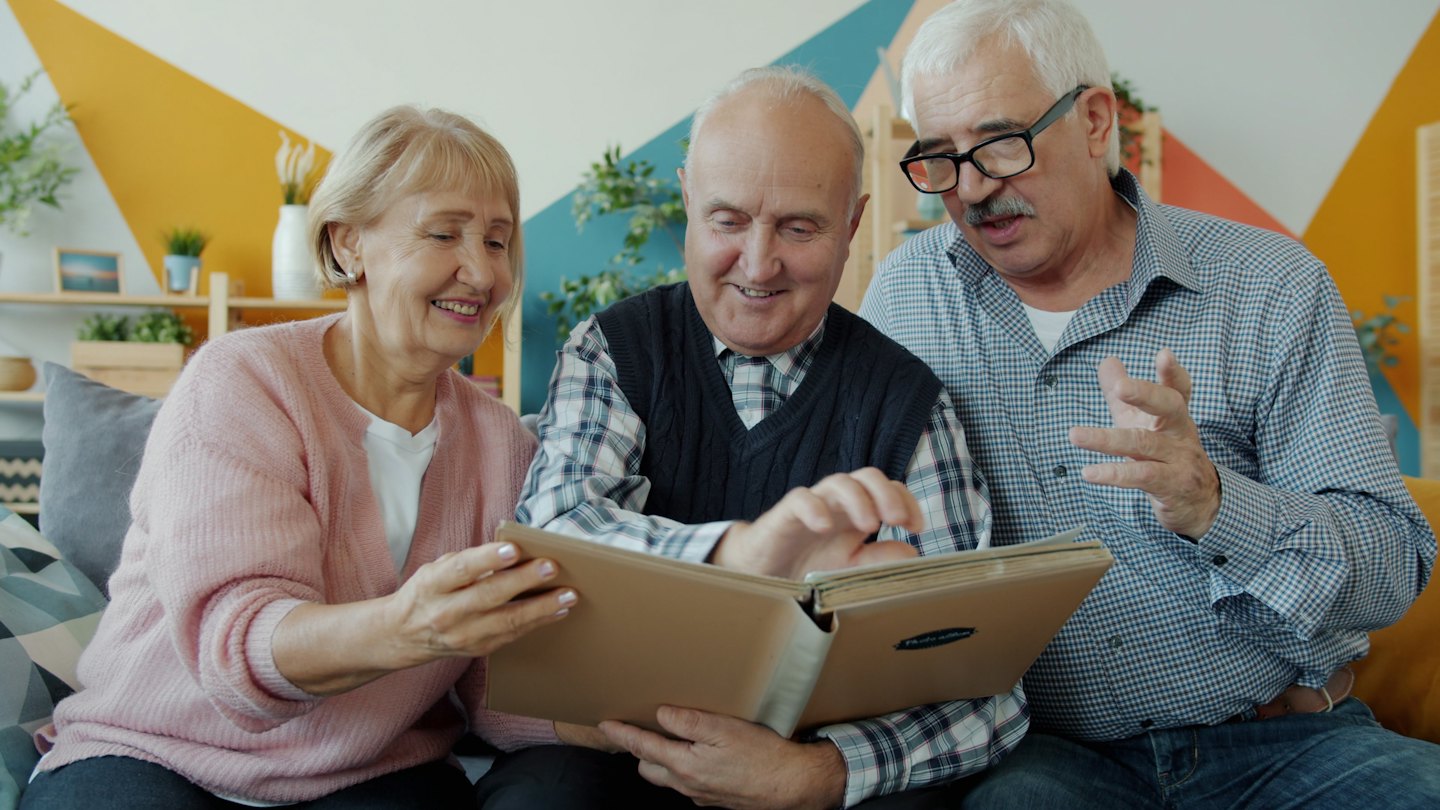 Three smiling adults look at a photo album on a sofa.