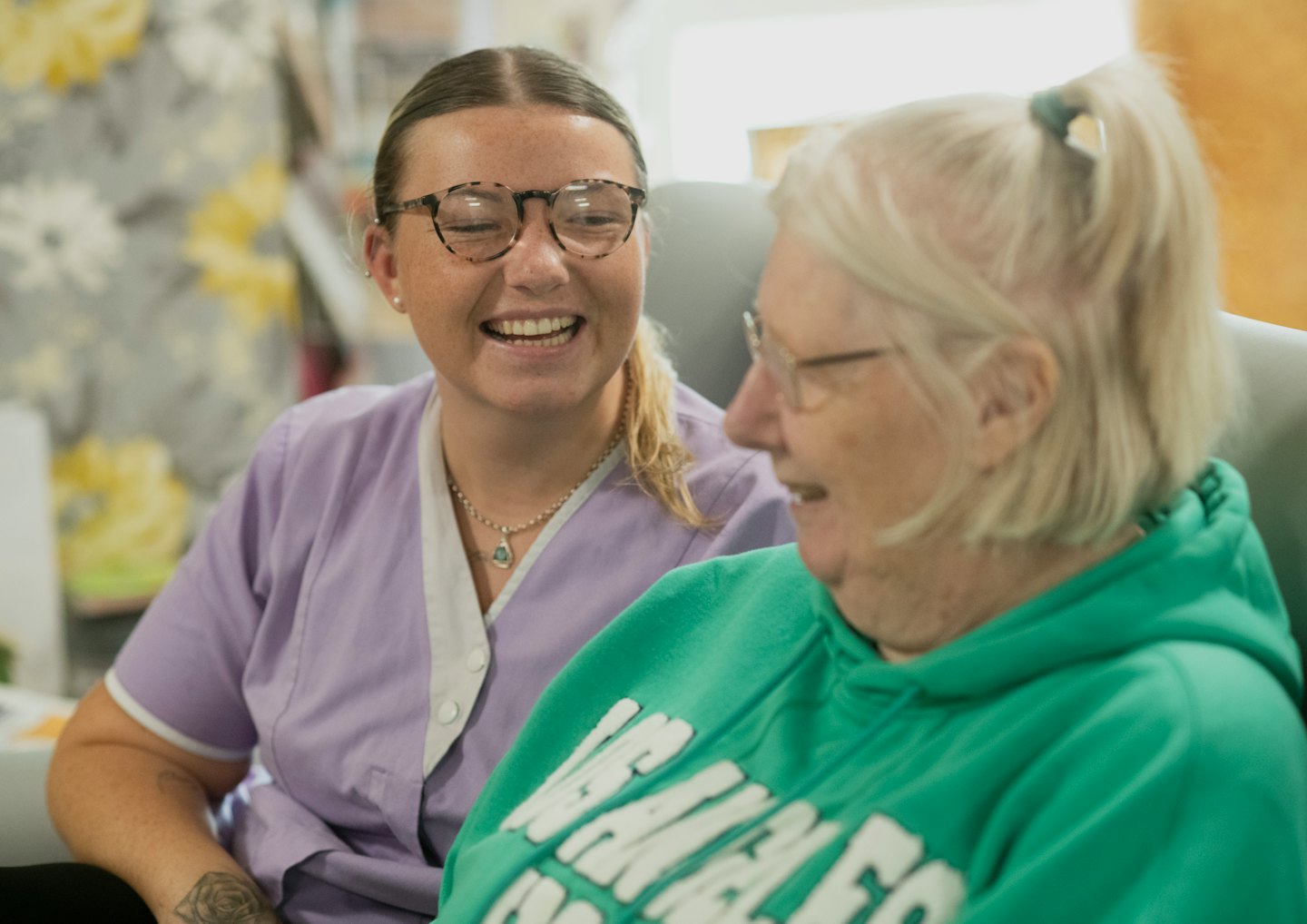 Caregiver in lavender uniform smiles with elderly woman.